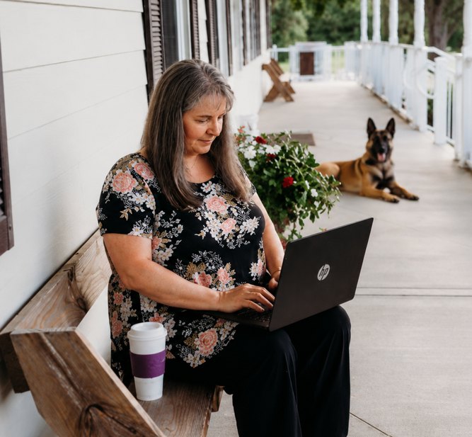 Linda Hubatsek working on a laptop on a sunny front porch, with a dog resting nearby.